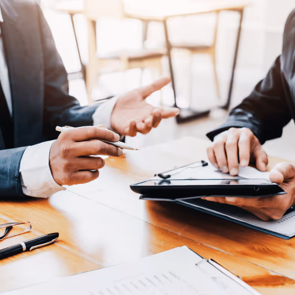 Dwóch mężczyzn w garniturach omawia dokumenty przy stole, jeden trzyma długopis, a drugi korzysta z tabletu.