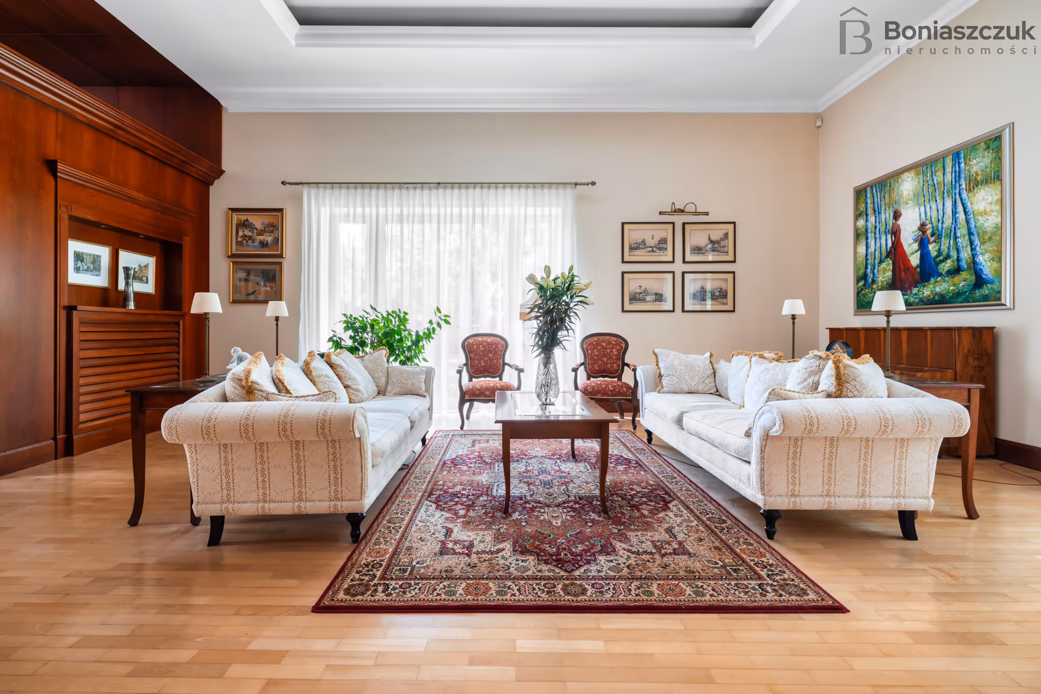 Przestronny salon z dwoma kremowymi sofami, czerwonymi krzesłami, drewnianym stolikiem i dekoracyjnym czerwonym dywanem.