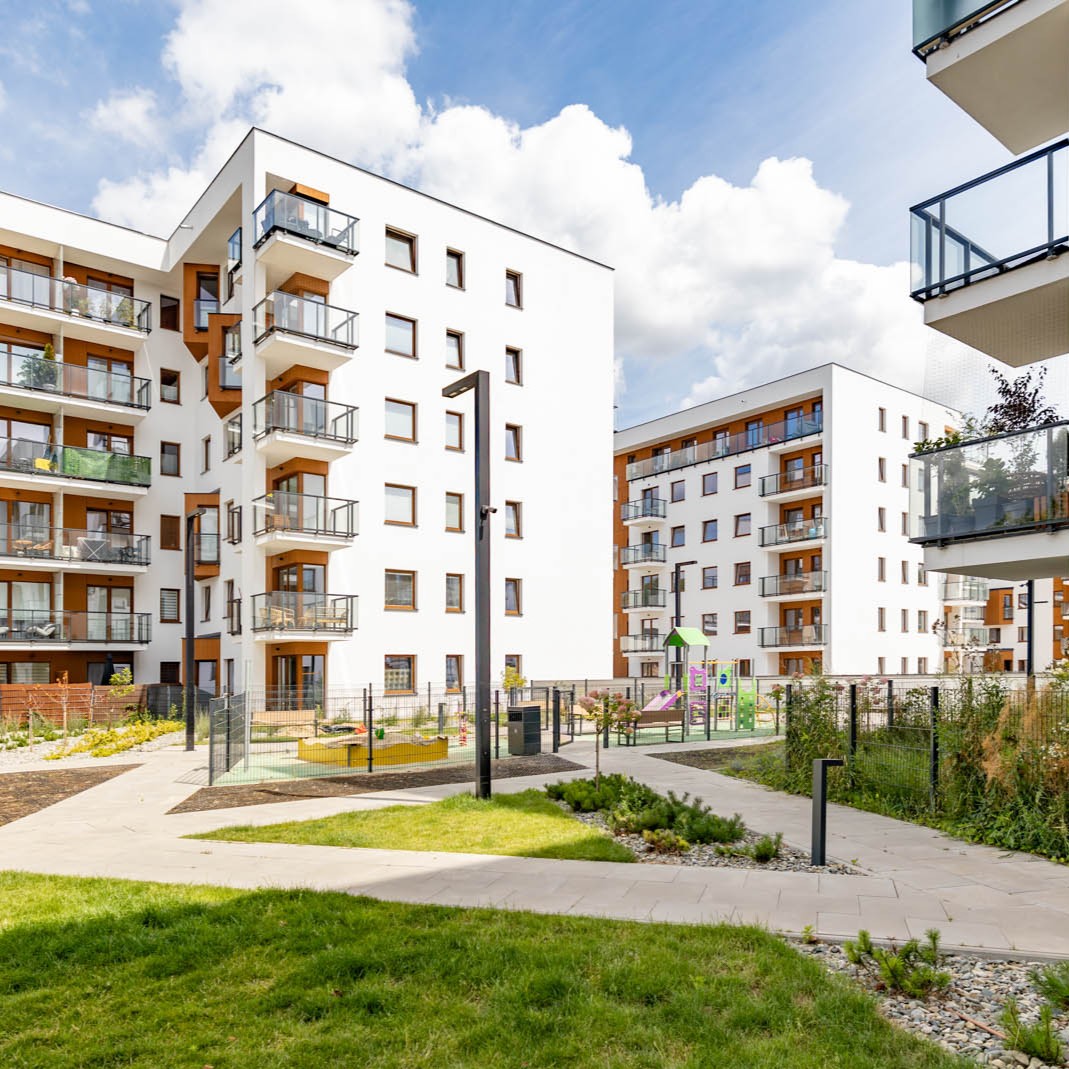 Nowoczesne osiedle mieszkaniowe z białymi budynkami, balkonami, placem zabaw i zielonym podwórkiem.