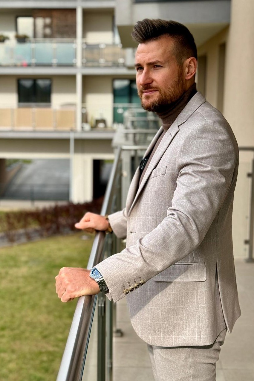 Mężczyzna w garniturze opartym o balustradę balkonu na tle budynków mieszkalnych.