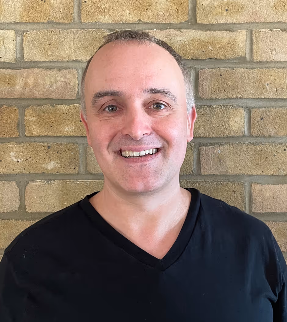 Smiling middle-aged man with short hair wearing a black V-neck shirt standing in front of a brick wall.