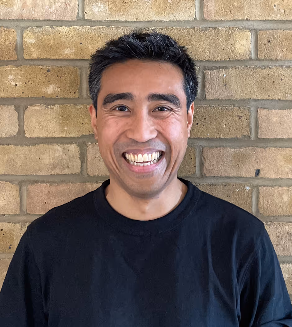 Smiling man with short black and gray hair wearing a black shirt standing in front of a brick wall.