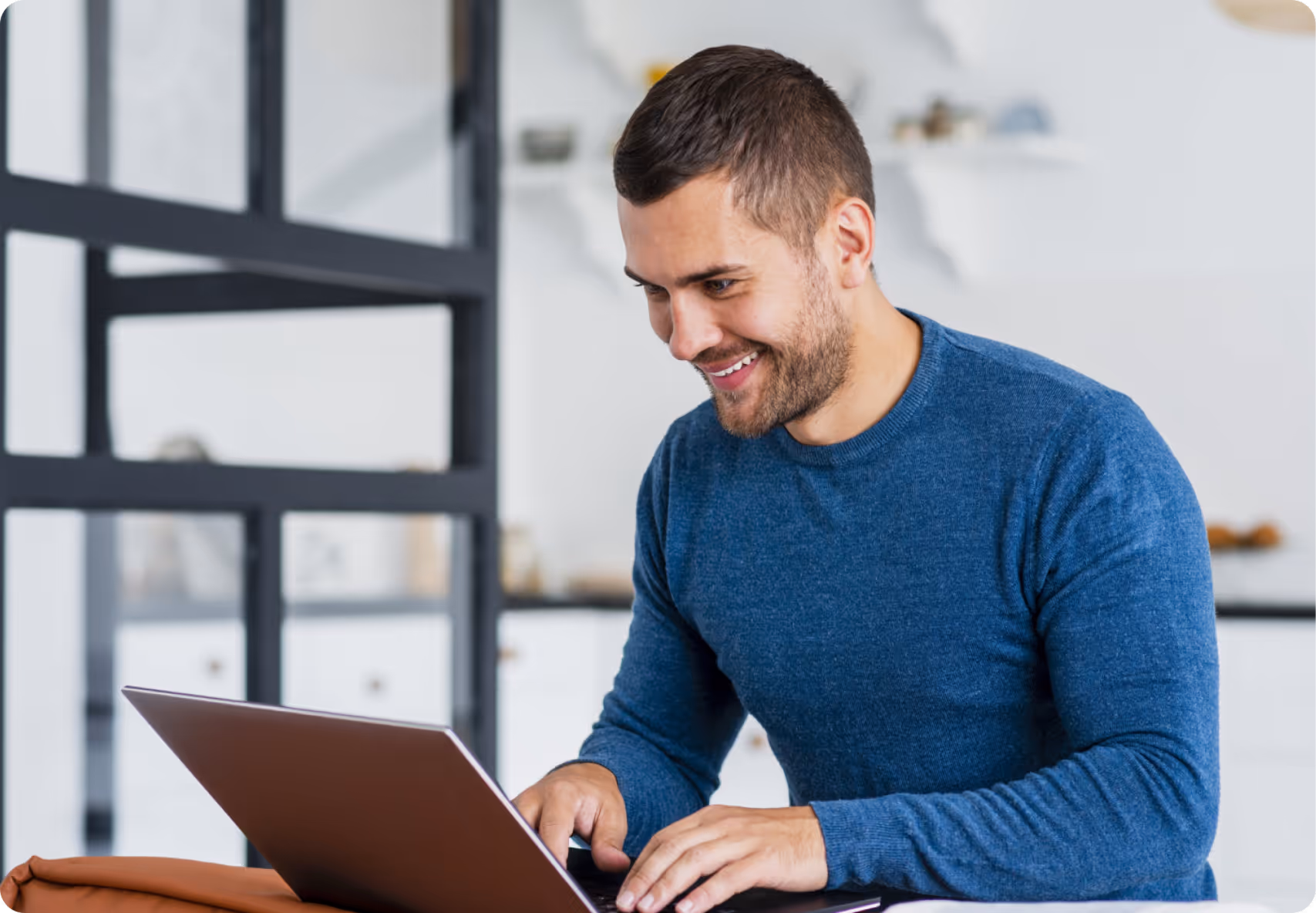 Smiling man in a blue sweater typing on a laptop in a modern room.