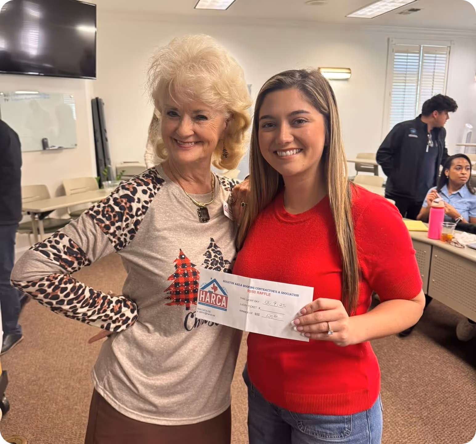 Two smiling women, one in a leopard print Christmas sweater and the other in a red sweater, holding a HARCA raffle check.