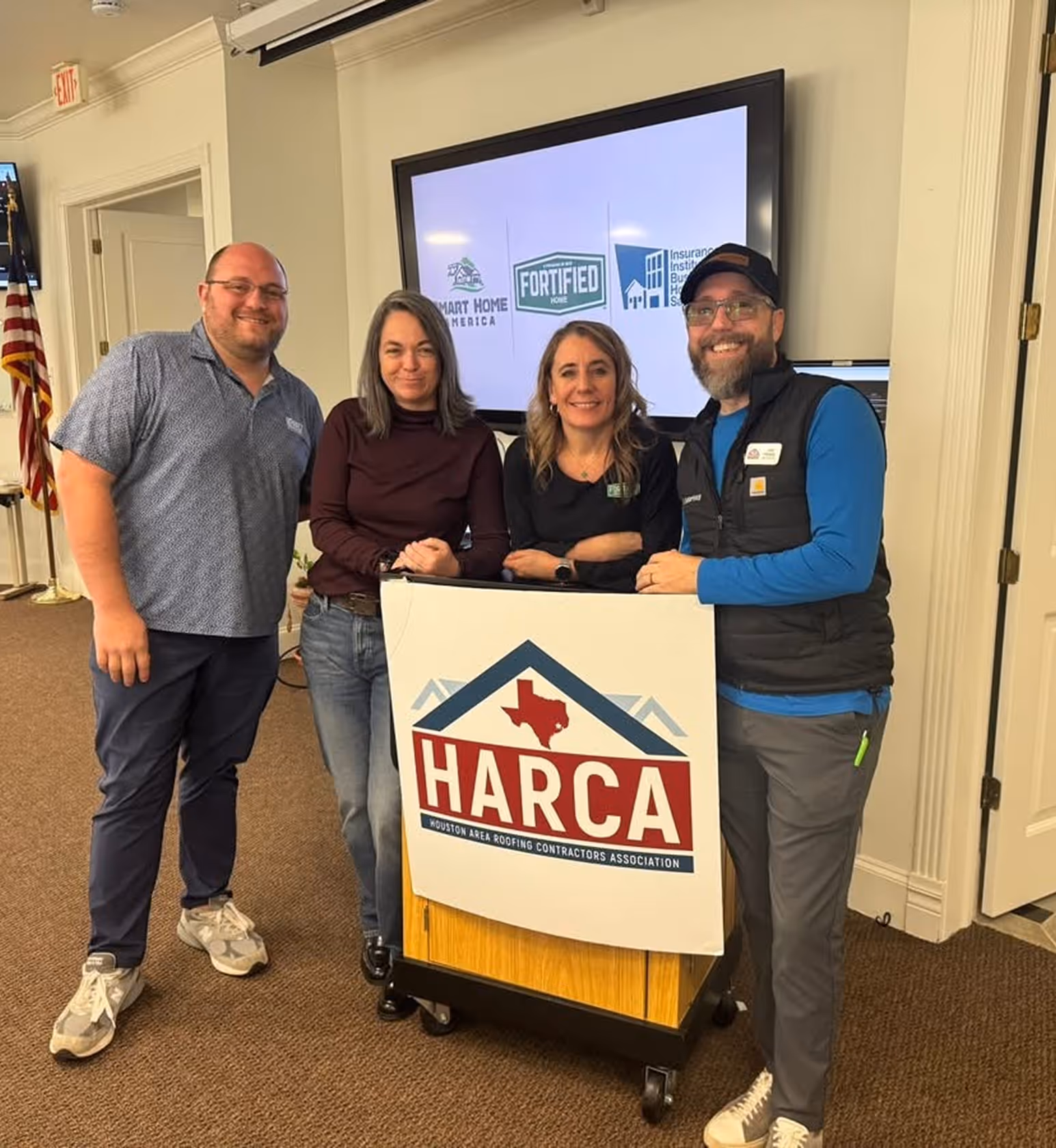 Four people standing indoors around a podium with a HARCA logo, smiling, with a screen behind displaying Smart Home America, Fortified Home, and Insurance Institute logos.