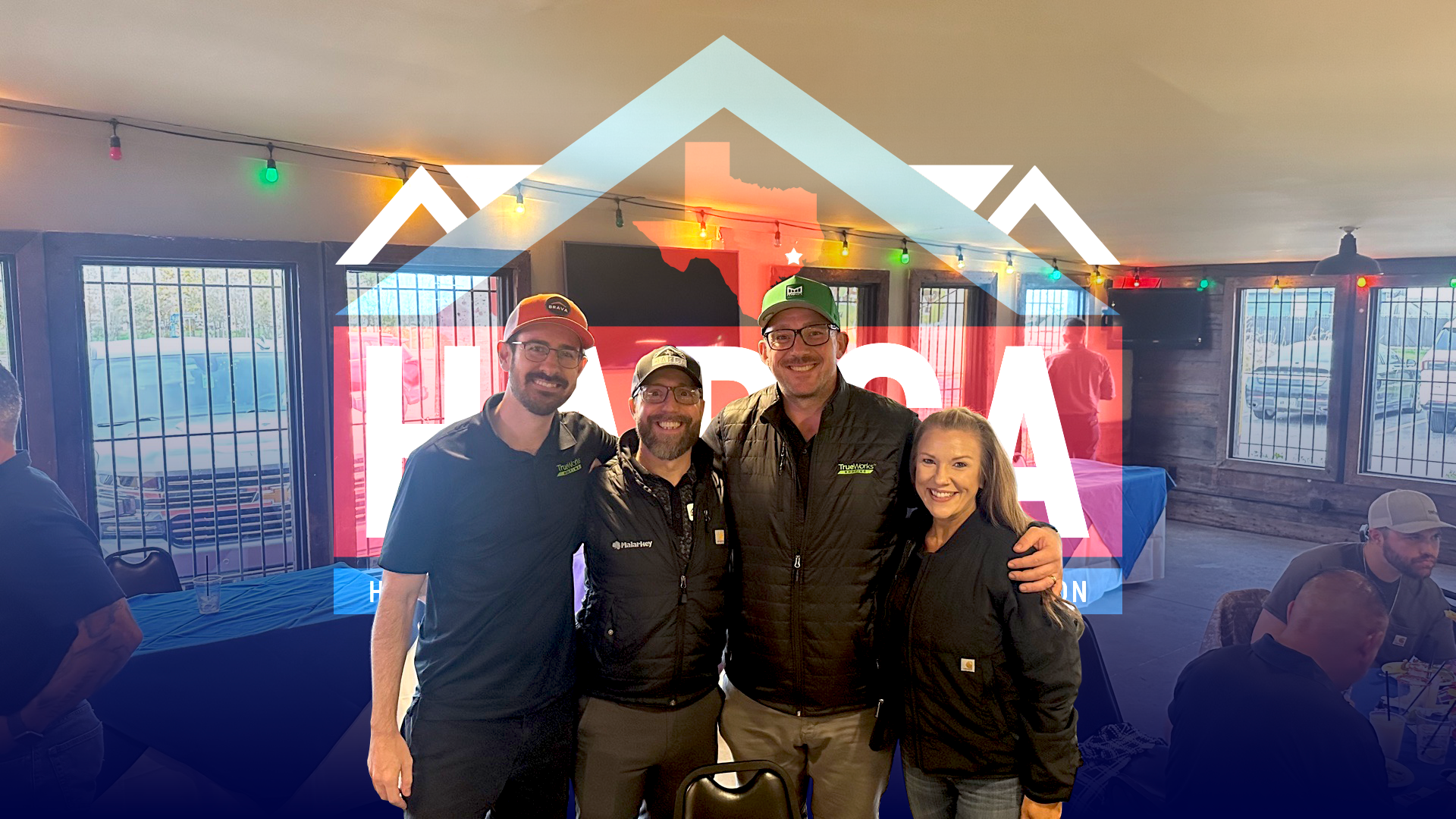 Four smiling people wearing black jackets standing indoors with string lights and barred windows in the background.