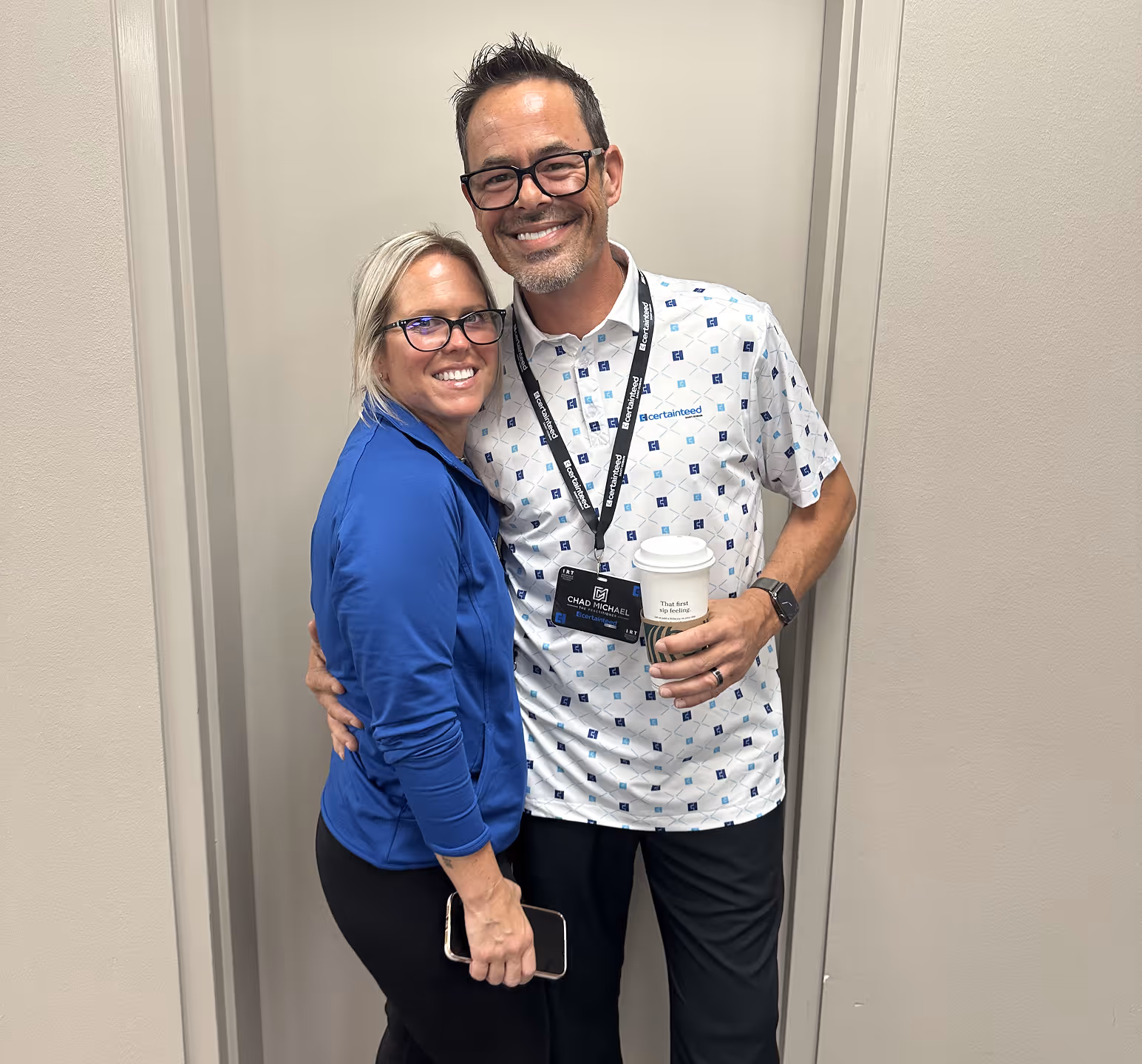 Smiling man and woman posing together, the man holding a coffee cup and wearing a work badge and patterned shirt.