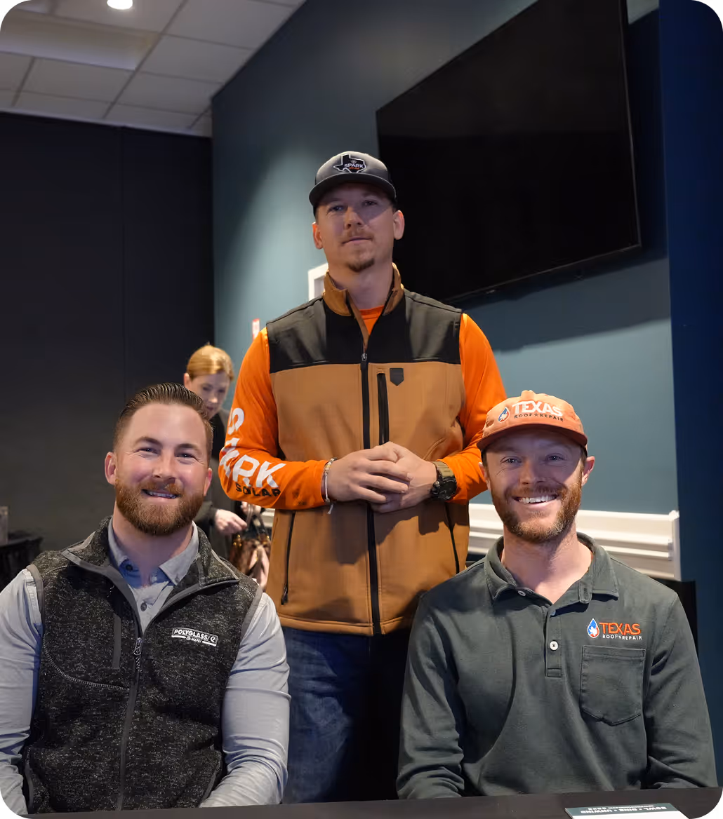 Three men posing indoors, two seated and one standing behind them, all wearing casual workwear with company logos.