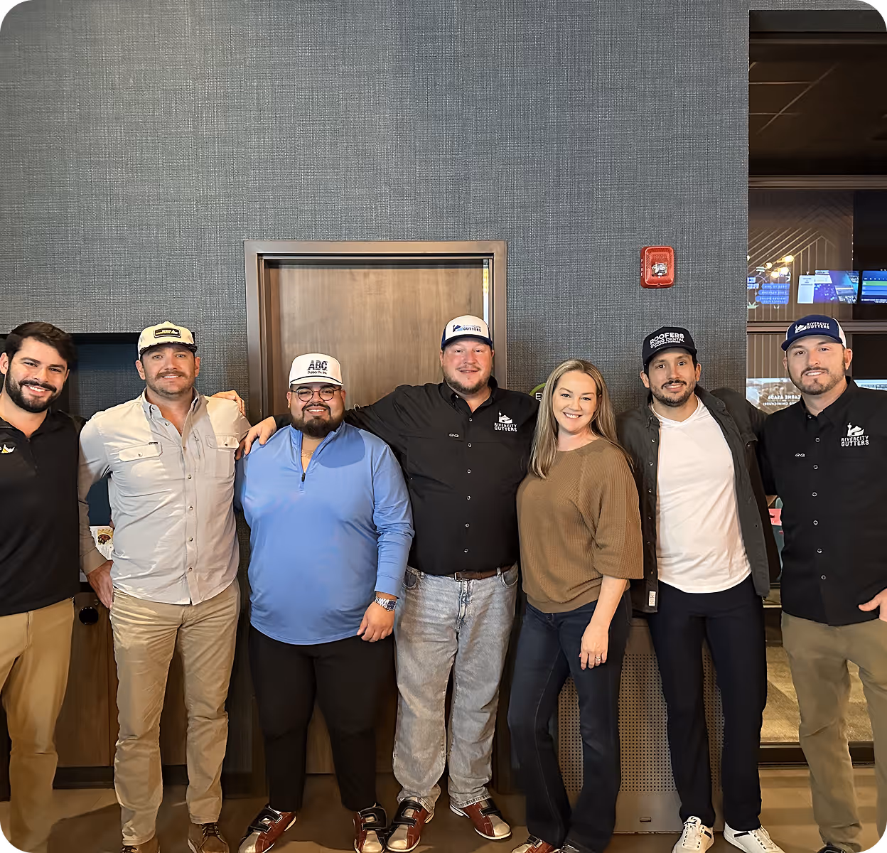 Group photo of six men and one woman standing indoors against a textured wall, smiling and casually dressed.