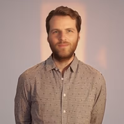 Homme avec barbe et cheveux courts portant une chemise grise à motifs, regardant vers l'avant avec un léger sourire.