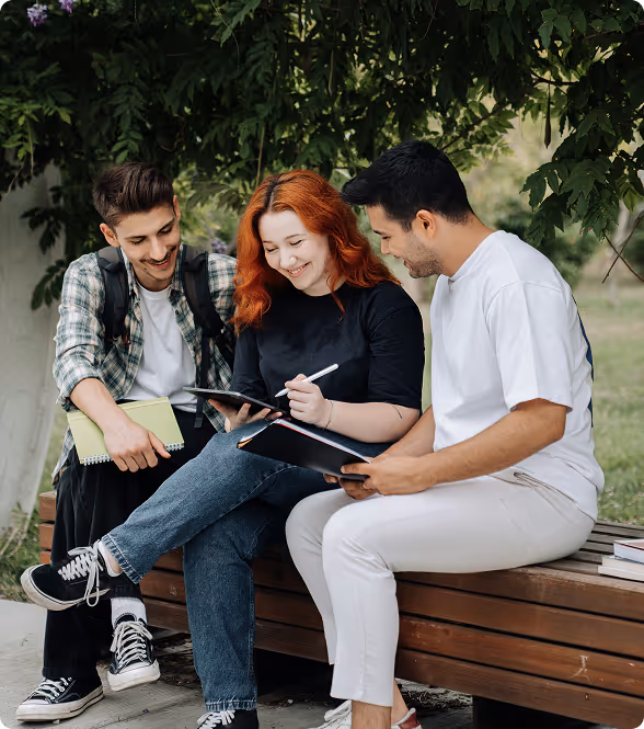 Trois étudiants assis sur un banc extérieur, souriants et collaborant en regardant des notes et une tablette.