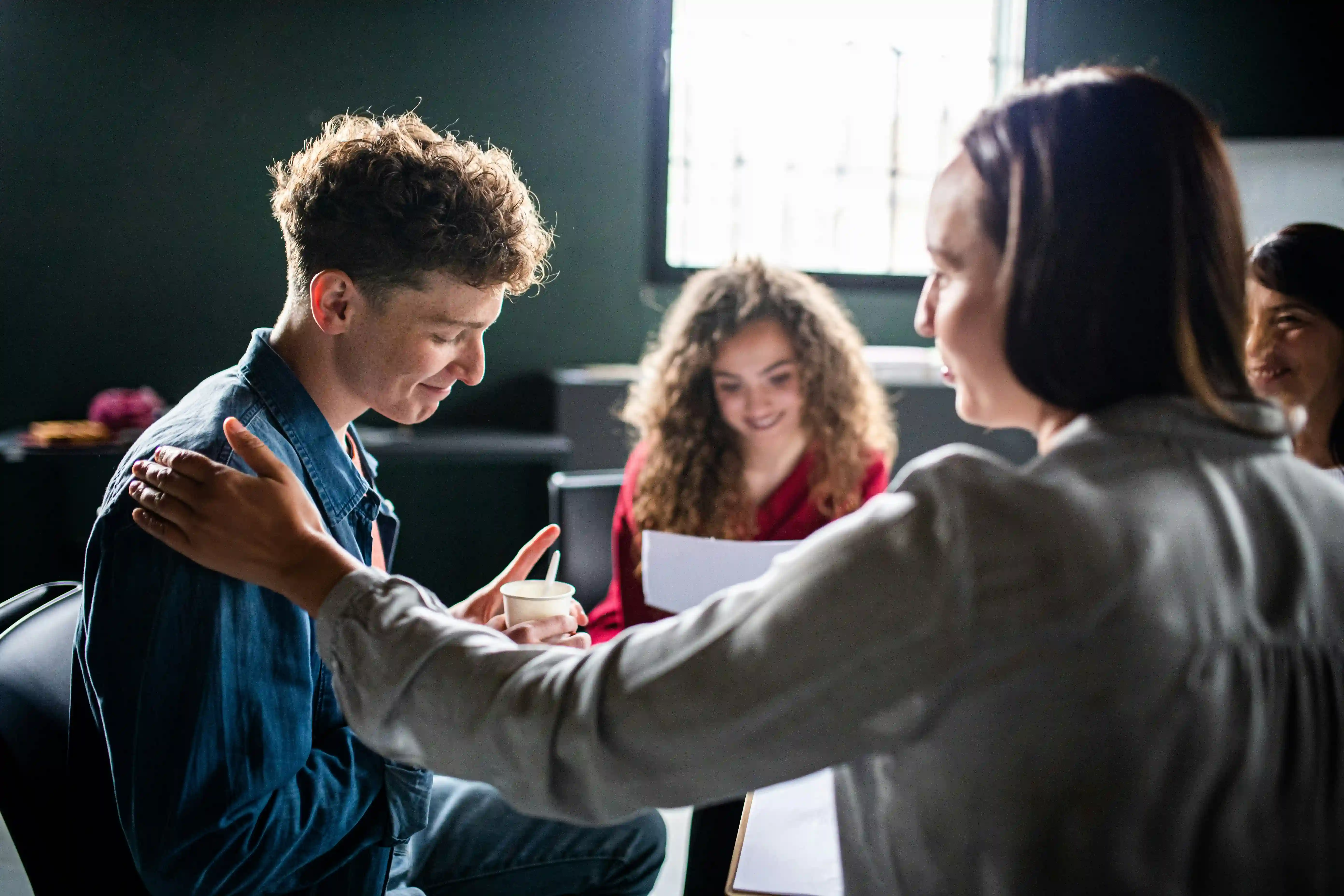 Jeunes adultes en discussion positive dans une pièce lumineuse, une femme pose la main sur l'épaule d'un homme souriant.