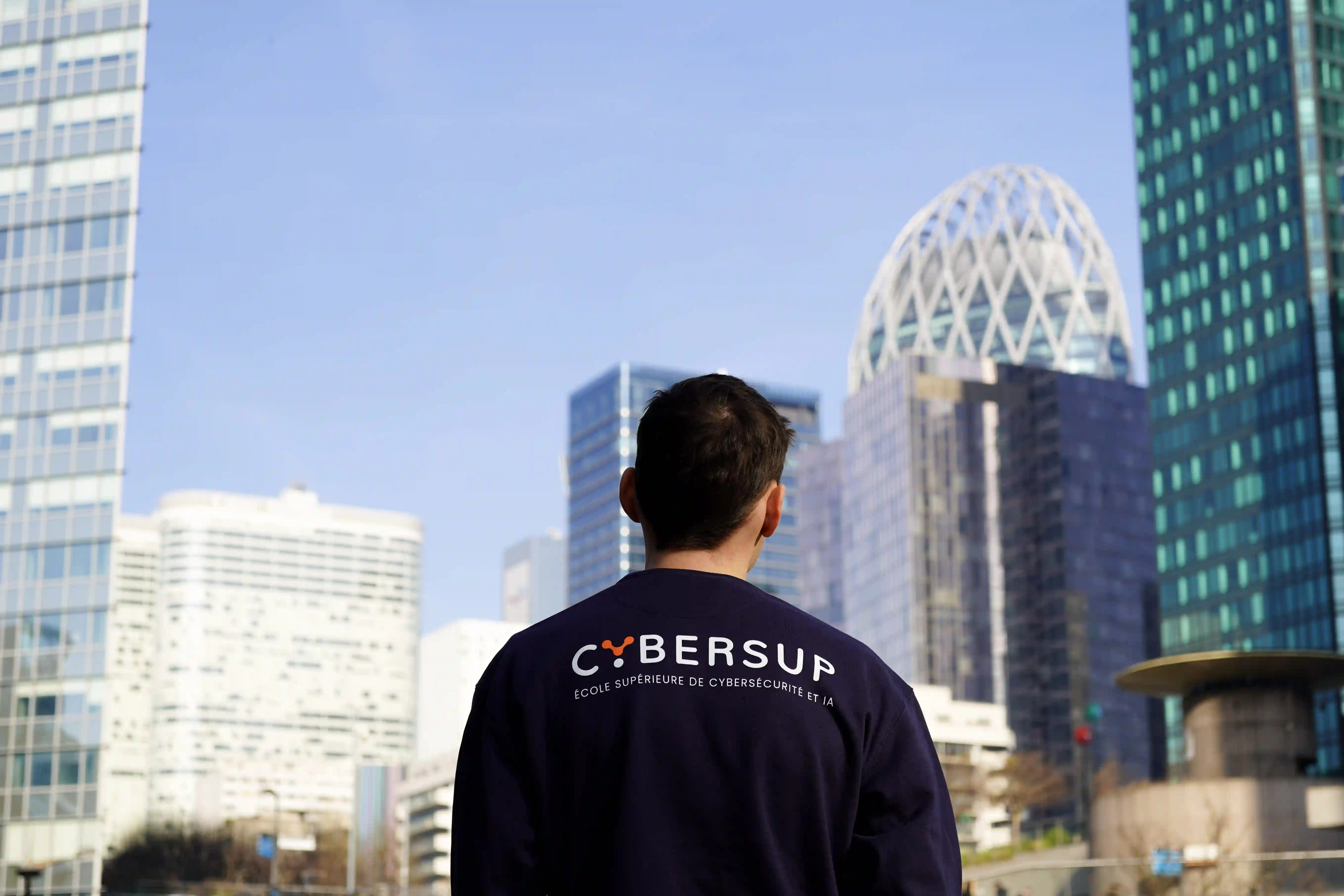 Un homme vu de dos porte un sweat-shirt avec le logo Cybersup devant des immeubles modernes ensoleillés.