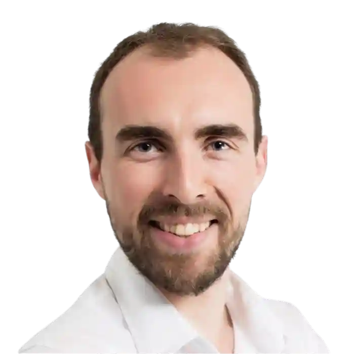 Homme avec barbe et cheveux courts portant une chemise grise à motifs, regardant vers l'avant avec un léger sourire.