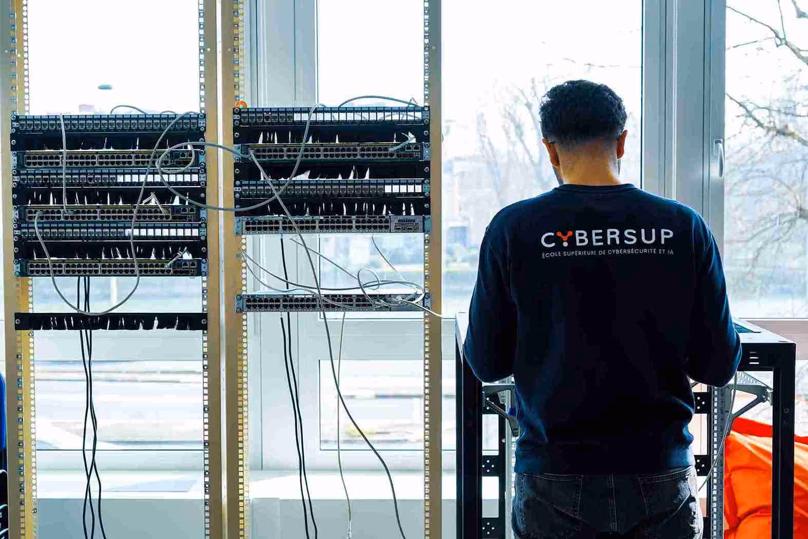 Personne de dos portant un sweat-shirt Cybersup travaillant devant des racks de câblage réseau près d'une fenêtre.