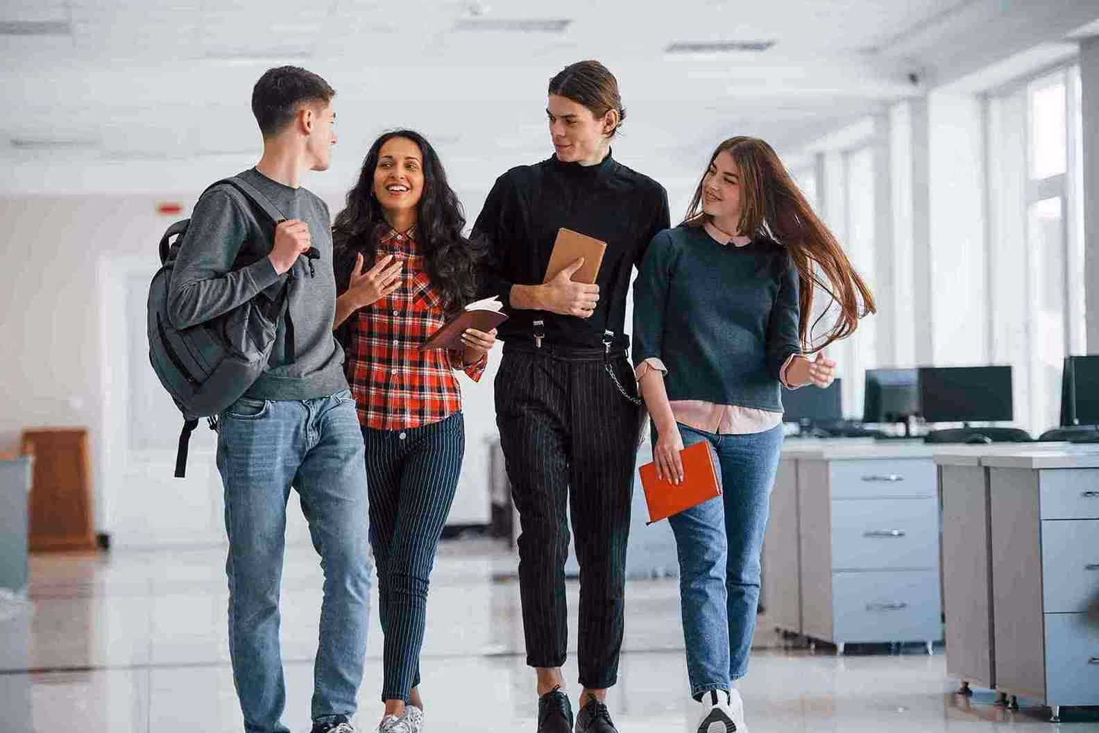 Quatre étudiants marchant ensemble dans un couloir d'école en discutant et souriant, avec des livres et un sac à dos.