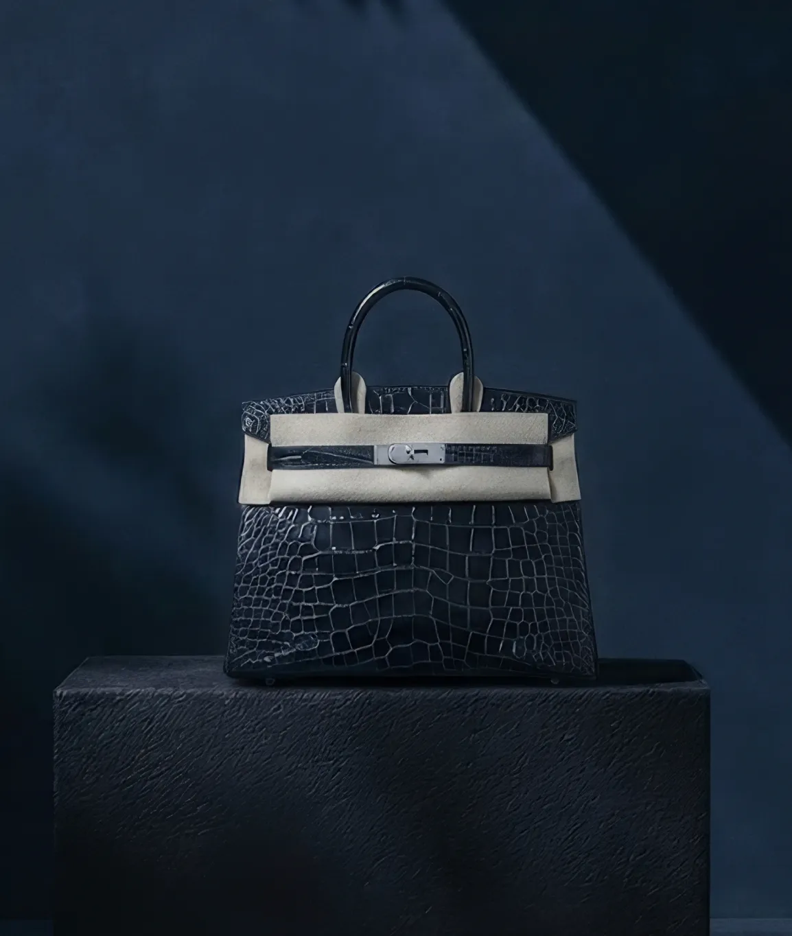 Black crocodile leather handbag with silver hardware placed on a dark textured pedestal against a dark blue background.