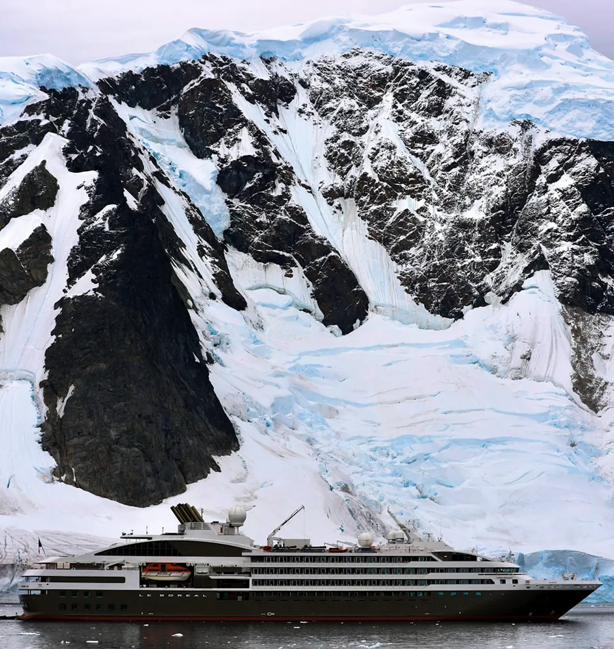 Cruise ship sailing beside massive snow-covered Antarctic cliffs.