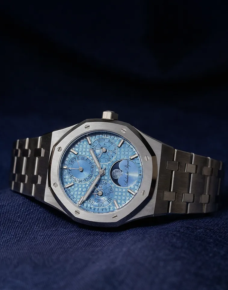 Silver wristwatch with a blue textured dial displaying time, date, day, month, and moon phase on a dark blue fabric background.