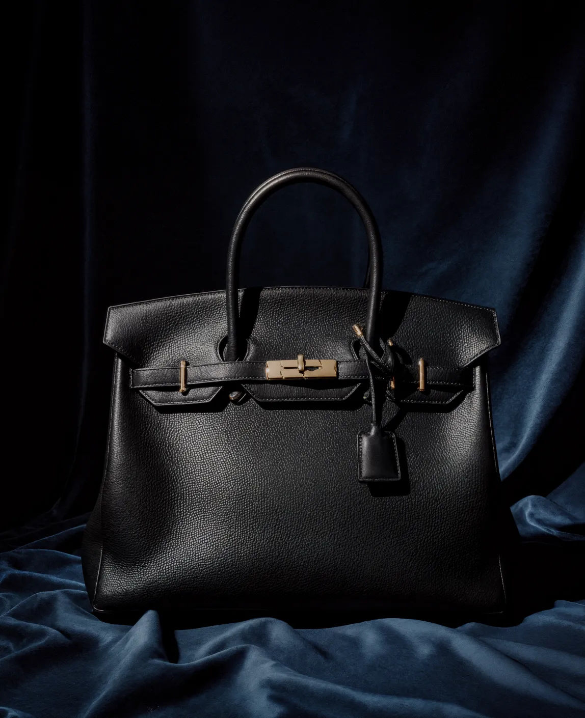Black leather handbag with gold hardware on a dark blue fabric background.