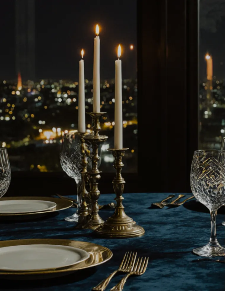 Elegant dining table with three lit white candles in brass holders, crystal glasses, and white plates set on a dark blue tablecloth with blurred city lights in the background.