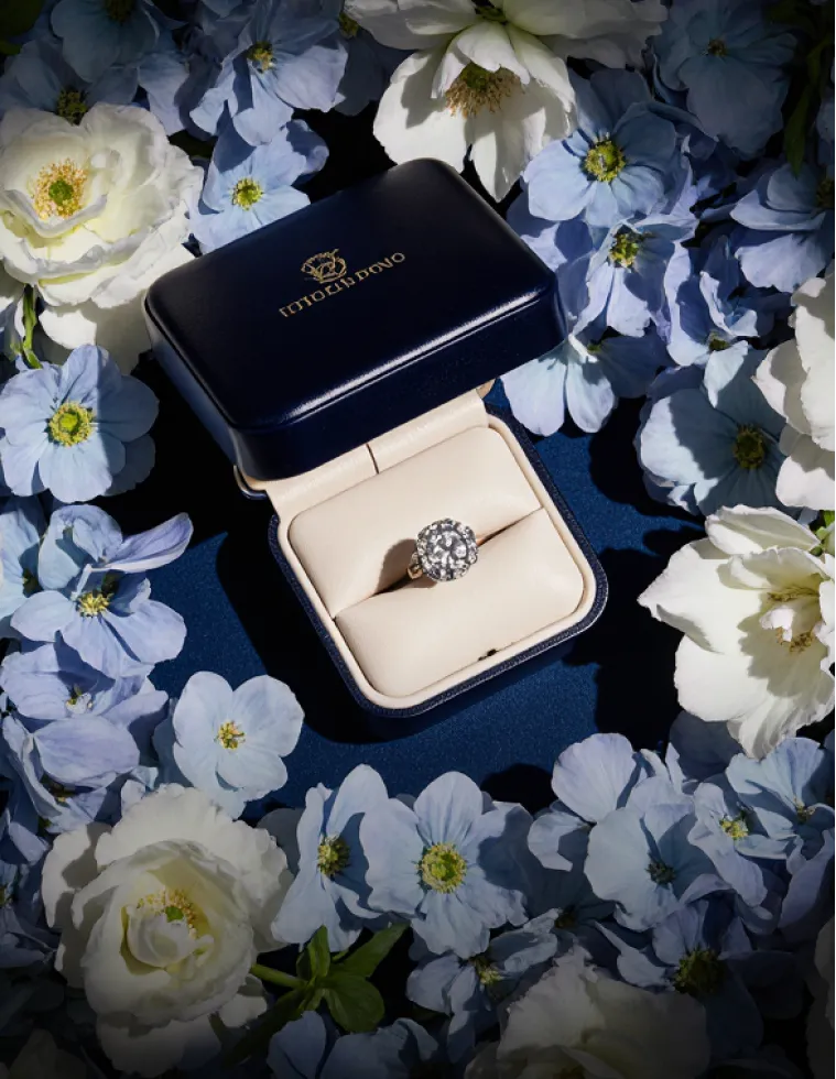 Diamond ring in an open blue and cream ring box surrounded by white and light blue flowers.