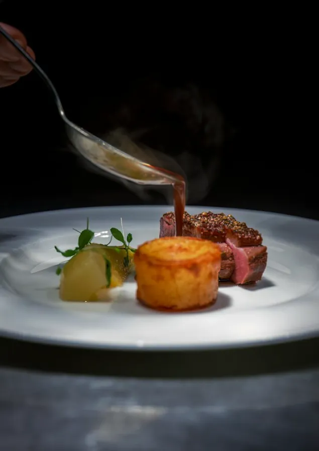 Hand pouring sauce from a spoon onto a plated gourmet dish with seared meat, a golden potato stack, and garnished vegetables on a white plate.