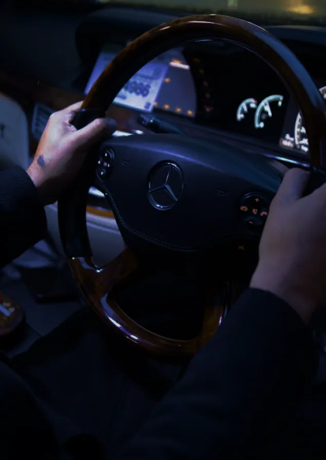 Person holding a wood-trimmed Mercedes-Benz steering wheel inside a car at night.