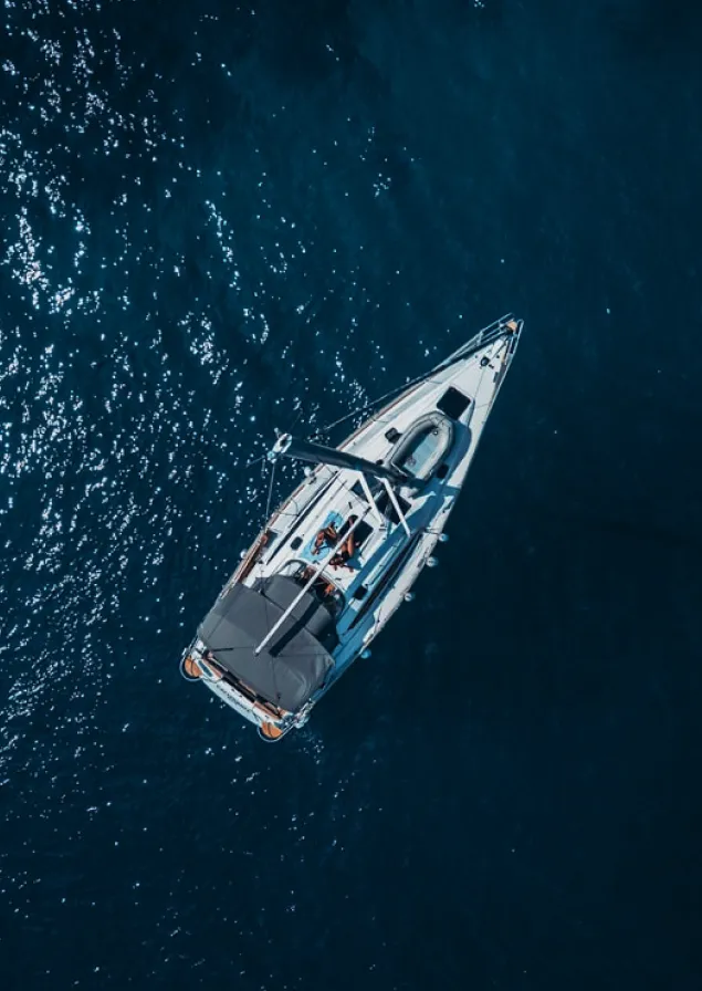 Aerial view of a sailboat with a person lying on the deck, floating on deep blue water with sunlight reflections.