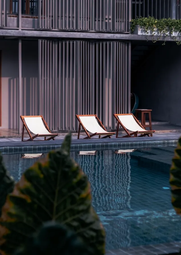 Three wooden lounge chairs with white fabric seats beside a modern swimming pool with large green leaves partially visible in the foreground.