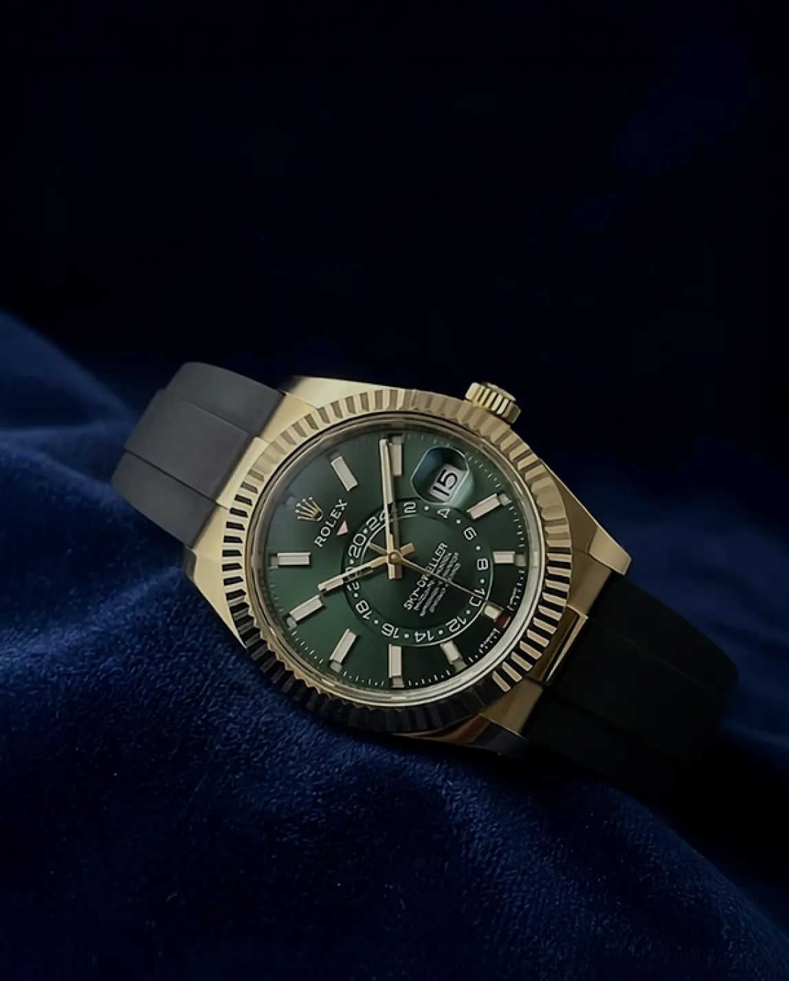 Rolex Sky-Dweller watch with green dial, gold bezel, and black rubber strap on dark blue fabric.