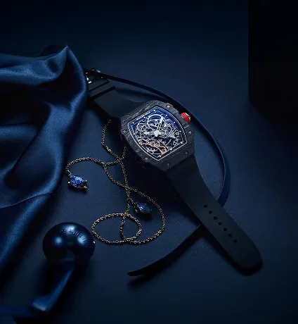 Luxury black wristwatch with visible inner mechanical parts, placed on a dark blue surface with a blue silk fabric, a gold chain with blue gemstone pendants, and a black key-shaped object.