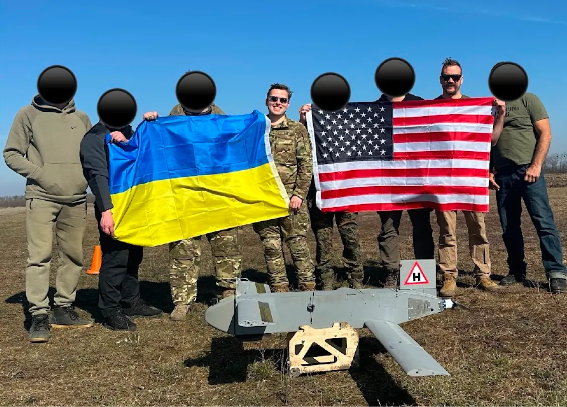Group of seven people standing outdoors on grass holding Ukrainian and American flags behind a small gray drone on a wooden stand.