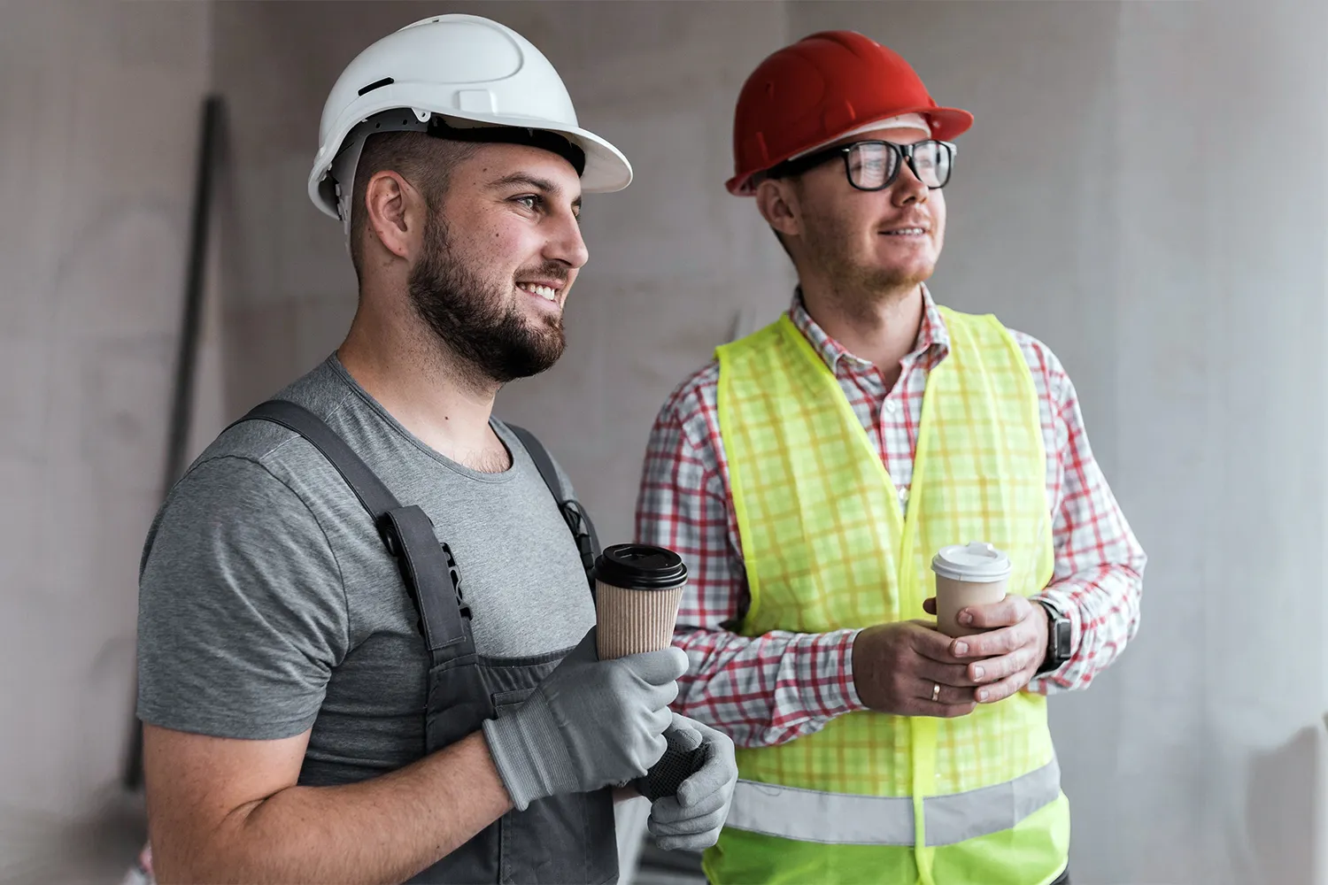 Zwei Bauarbeiter mit Schutzhelmen und Arbeitskleidung halten Kaffeebecher und schauen lächelnd in die Ferne.