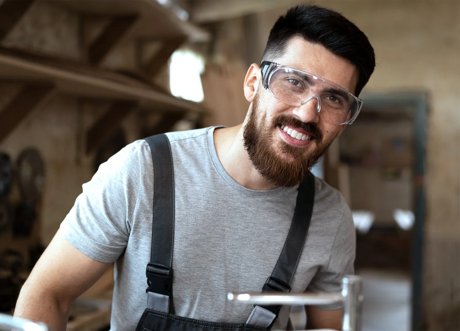 Junger Handwerker mit Schutzbrille und grau-schwarzer Arbeitskleidung lächelt in einer Werkstatt. https://www.magnific.com/de
