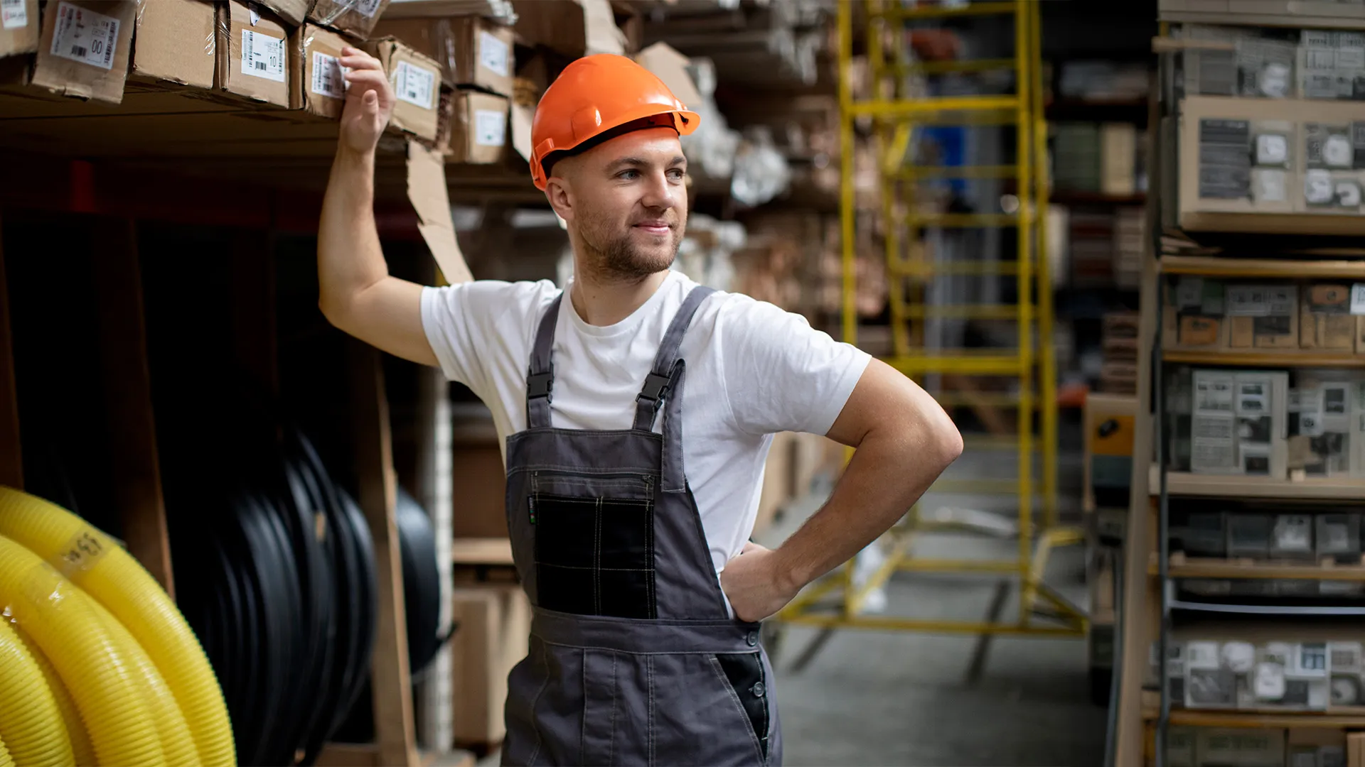 Arbeiter mit orangenem Schutzhelm und grauer Arbeitskleidung steht in einem Lager und hält sich eine Hand an die Hüfte. https://www.magnific.com/de