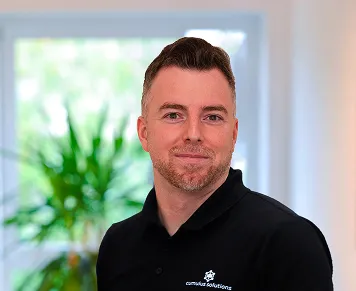 Smiling man with short hair wearing a black polo shirt standing indoors with a blurred window and green plant in the background.