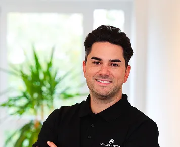 Smiling man with dark hair wearing a black polo shirt standing indoors with a green plant and window in the background.