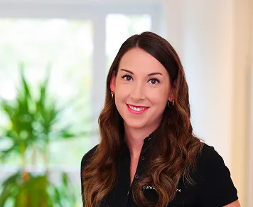 Smiling woman with long brown hair wearing a black top standing indoors with a plant blurred in the background.