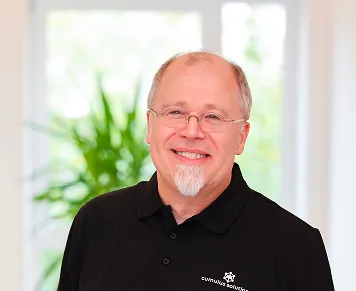 Smiling middle-aged man with glasses and goatee wearing a black polo shirt standing indoors with a blurred green plant in the background.