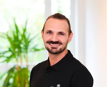 Smiling man with a beard and tied-back hair wearing a black polo shirt, standing indoors with green plants and bright windows in the background.