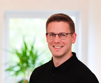 Smiling man with short brown hair and glasses wearing a black polo shirt in front of a blurred window with greenery.