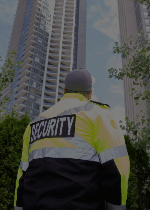 Security officer in a high-visibility jacket standing outdoors with tall buildings and trees in the background.