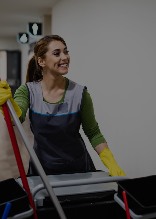 Smiling woman wearing yellow cleaning gloves and apron, pushing a cleaning cart with mop in a hallway.