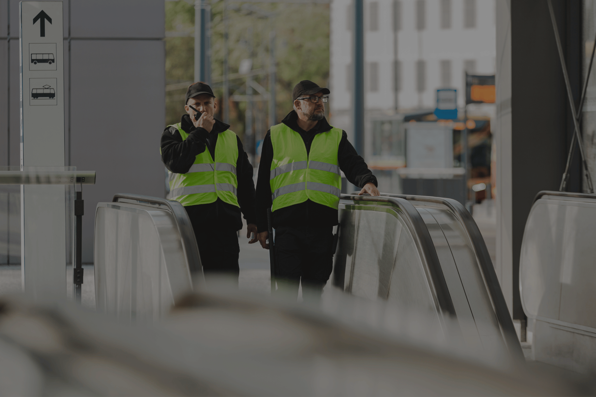 Two security guards wearing yellow reflective vests and black caps standing near escalators in a public transit area, one speaking into a walkie-talkie.