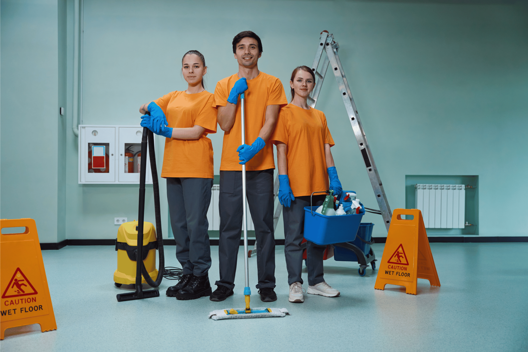Three cleaning workers in orange shirts and blue gloves standing with cleaning equipment, including a vacuum, mop, and bucket with supplies, in a room with caution wet floor signs.