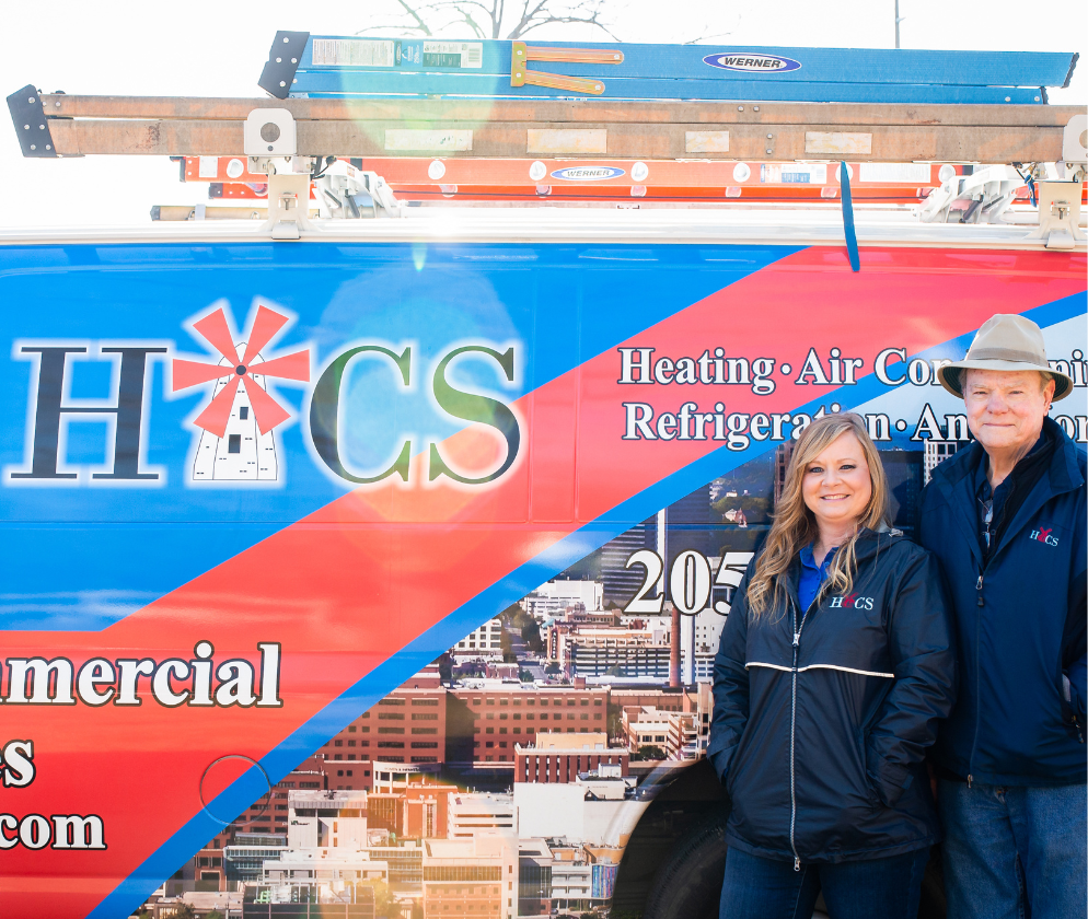 Two smiling people wearing black jackets stand next to a colorful service van with ladders on top, displaying a heating and air company logo.