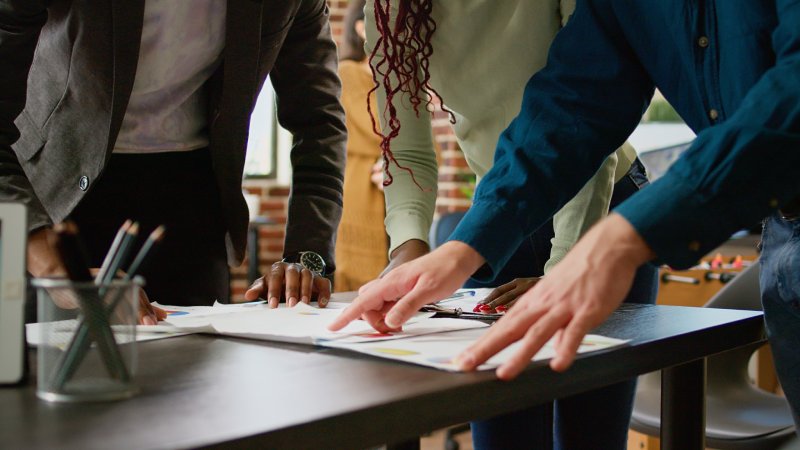 Three people collaboratively reviewing documents spread on a table in an office setting.
