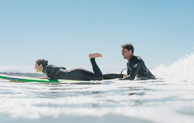 Un homme et une femme portant des combinaisons de plongée sur des planches de surf, attendant une vague dans l'océan. La femme est allongée sur sa planche tandis que l'homme est en train de ramer. Le ciel est bleu et dégagé