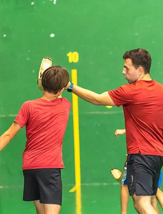 Un entraîneur de pelote basque, portant un t-shirt rouge, guide un jeune joueur en l'aidant à tenir sa raquette de chistera ou de pala contre un fronton vert